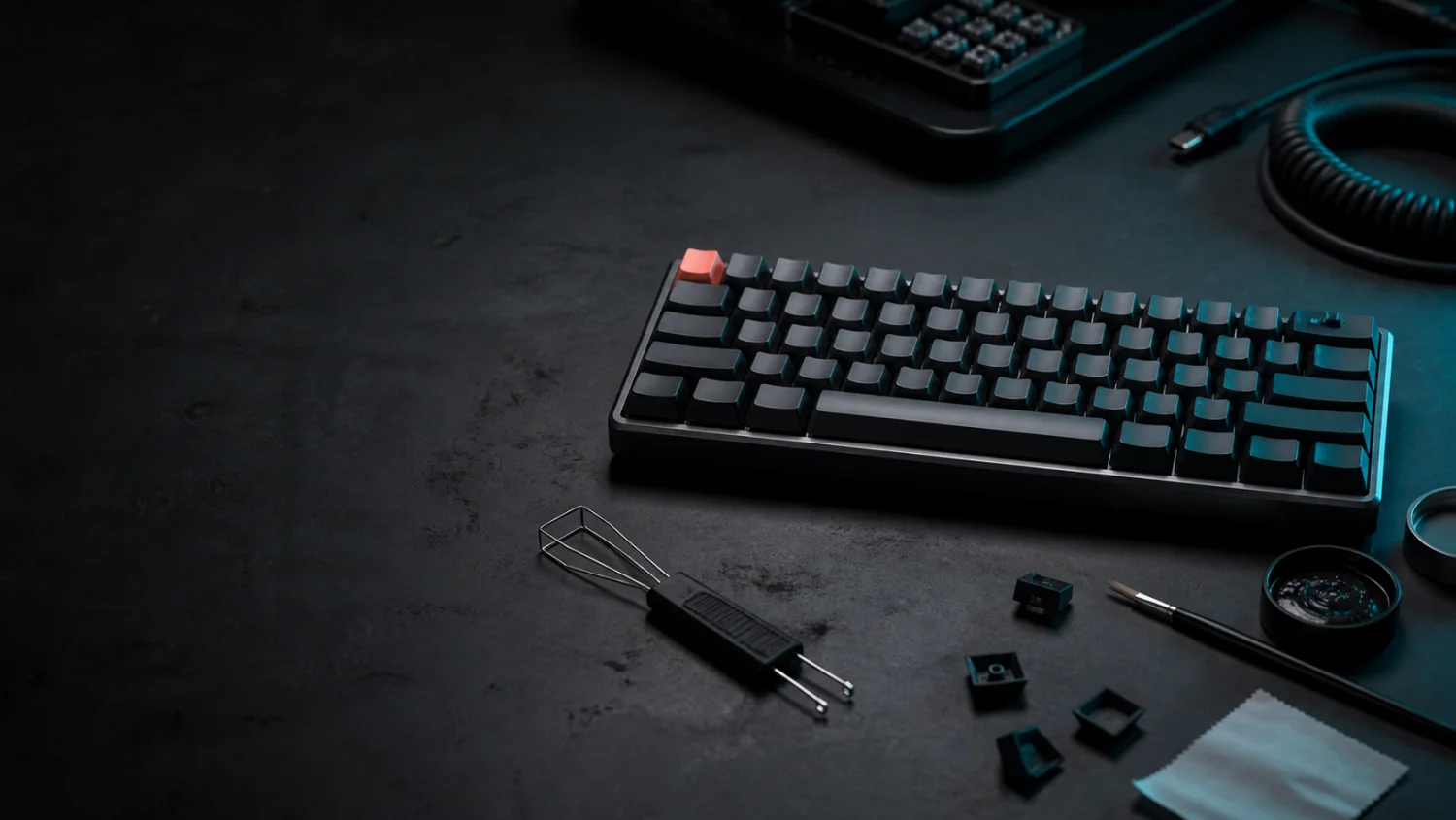 A mechanical keyboard workbench with loose keycaps, a keycap puller, a small brush, and a coiled cable